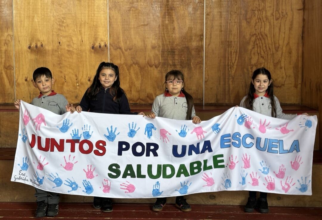 Cuatro estudiantes de párvulos sosteniendo un lienzo "Juntos por una escuela saludable" con huellas de manos pintadas